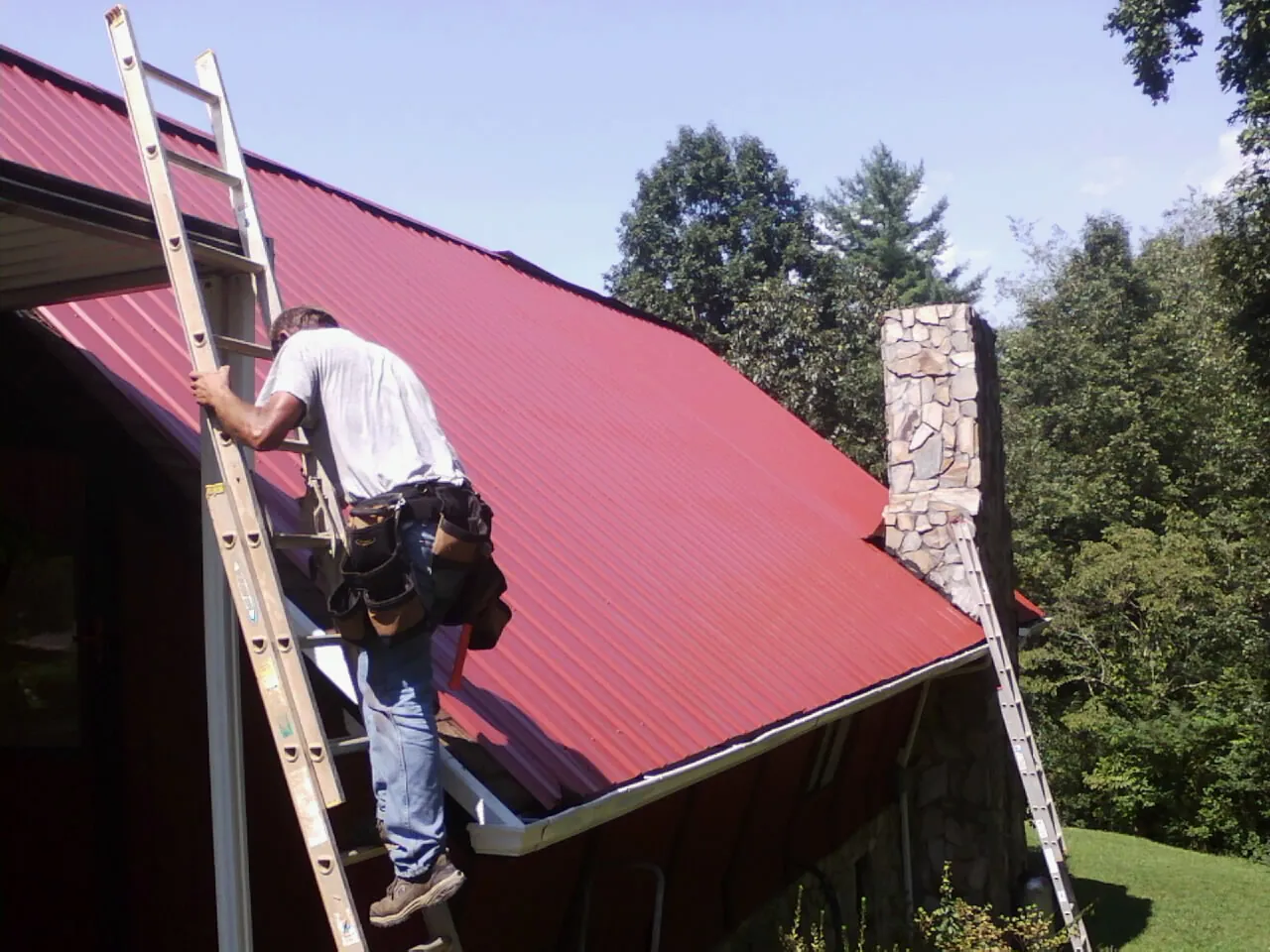 Roofer on a ladder beside a red metal roof and stone chimney — Soffit & Fascia Repair by Roofing Brothers in Lake Pleasant